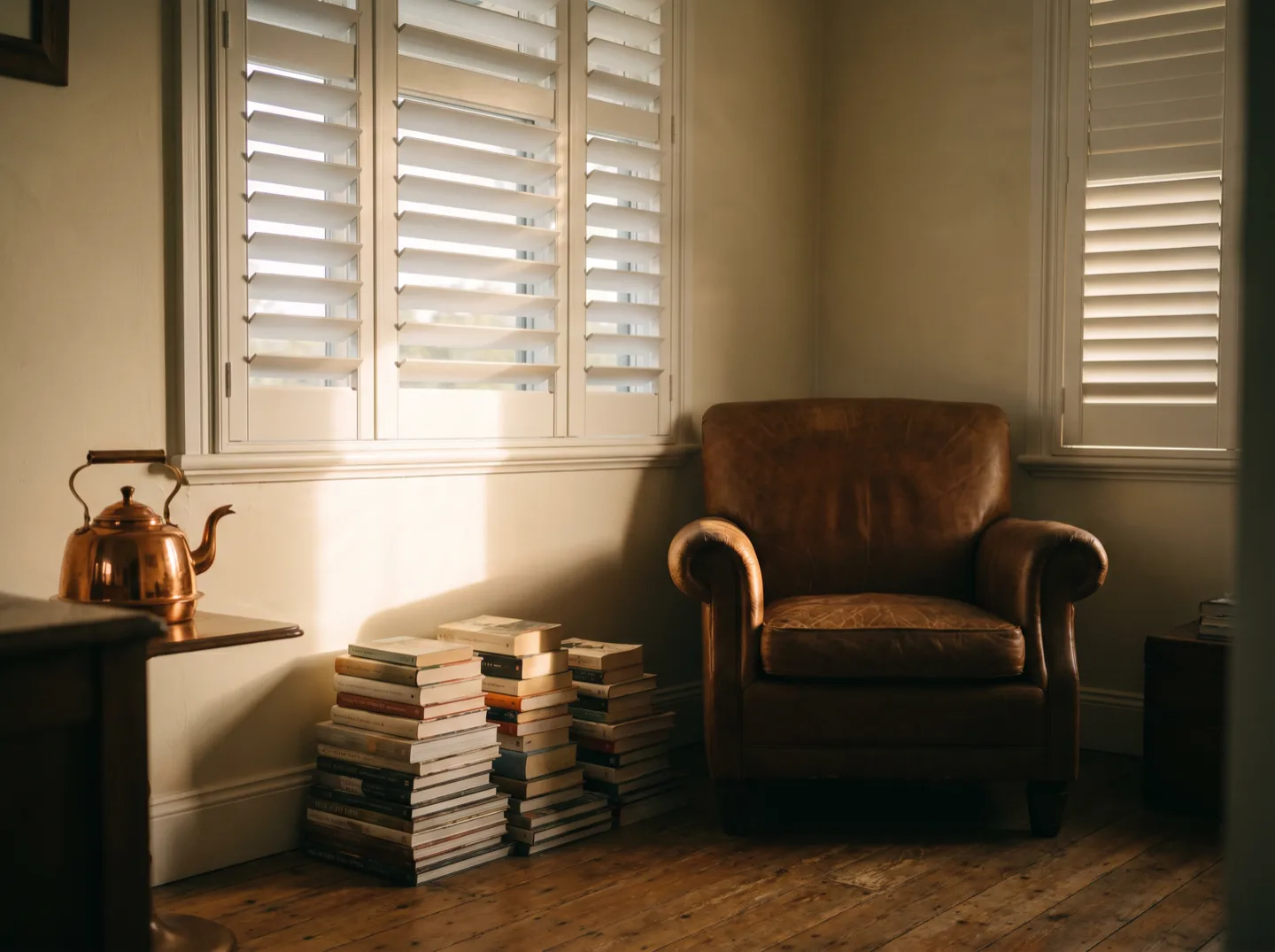 A front room with plantation shutters angled for privacy, reading chair and stacked books in warm light